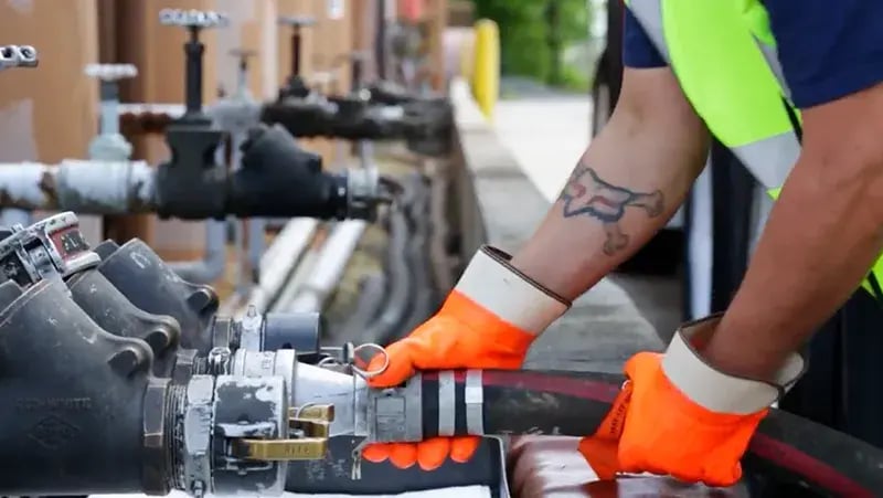 Worker in high-visibility gear connecting industrial hoses for fluid transfer at a fueling distribution station