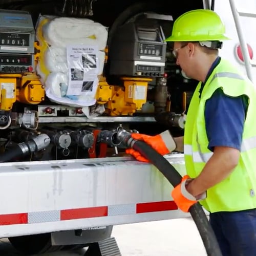 Worker in safety gear refueling a truck using a large hose at an industrial fueling station.